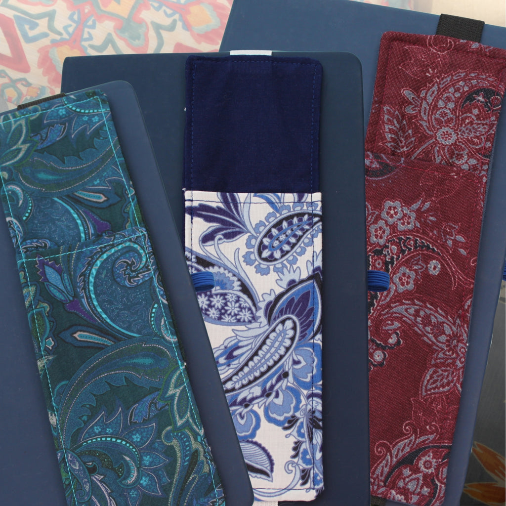 Three handmade A5 fabric notebook pen pockets fanned out on a blue notebook, made from pre-loved paisley fabrics in teal, blue and white, and burgundy — Dark Star Stitches Second Chapter collection