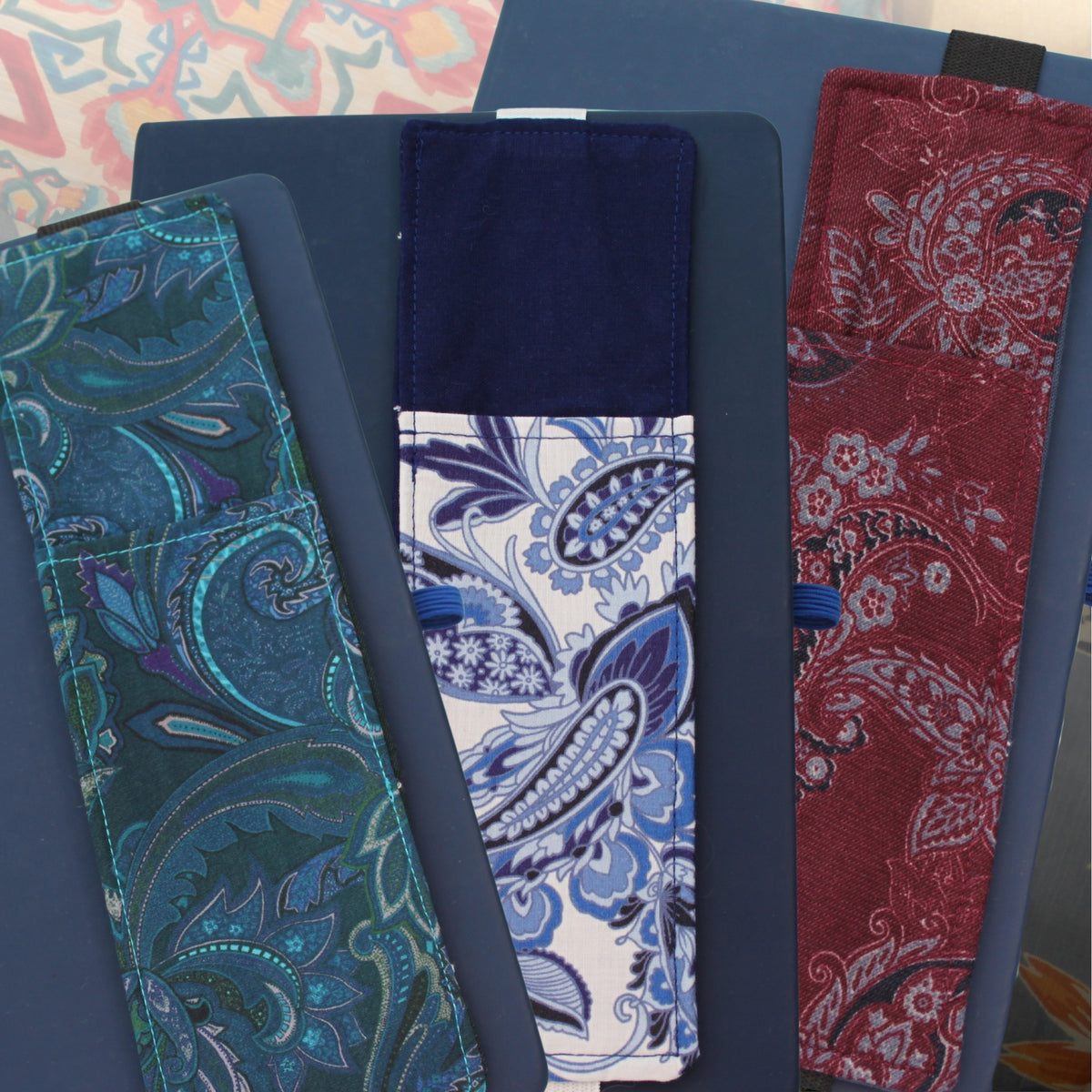 Three handmade A5 fabric notebook pen pockets fanned out on a blue notebook, made from pre-loved paisley fabrics in teal, blue and white, and burgundy — Dark Star Stitches Second Chapter collection